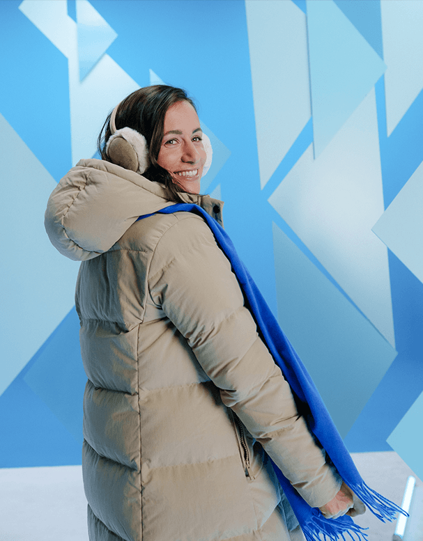 Frau in langer Daunenjacke und Ohrenschützern, trägt einen blauen Schal, steht vor einem blauen geometrischen Hintergrund.