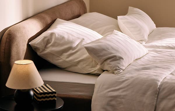 Gemütliches Bett mit weißen Kissen und Bettwäsche, daneben ein Nachttisch mit Lampe und Buchbox