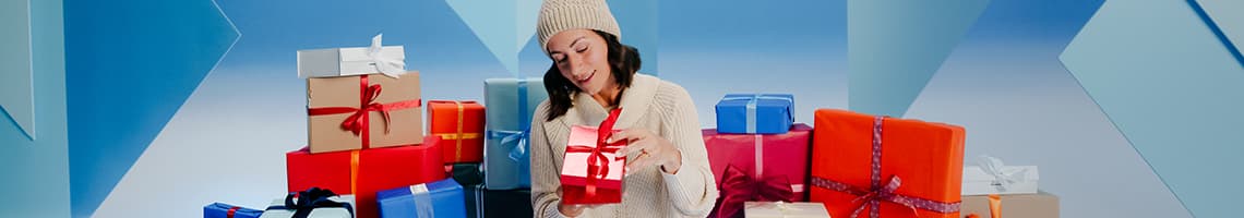 Frau in weißem Pullover und Mütze hält ein Geschenk, umgeben von vielen bunt verpackten Geschenken vor blauem Hintergrund