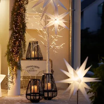 Beleuchtete Sternlampen und Laternen auf einer Terrasse mit Weihnachtsdeko und einem Pflanzwagen im Hintergrund