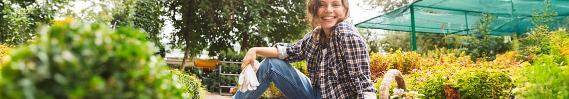 Person in kariertem Hemd und Jeans sitzt im Garten zwischen Pflanzen und Blumen, trägt weiße Handschuhe.