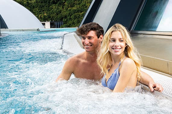 Paar sitzt entspannt im sprudelnden Außenpool, lehnt am Beckenrand mit moderner Architektur im Hintergrund