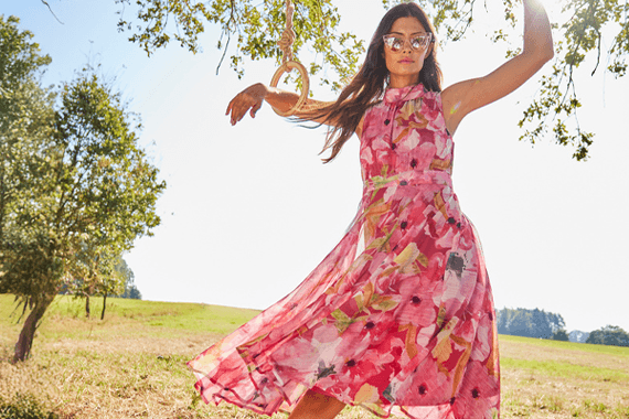 Frau im geblümten, knöchellangen Kleid steht auf einer Wiese und hält sich an einem Ast mit Schaukel fest