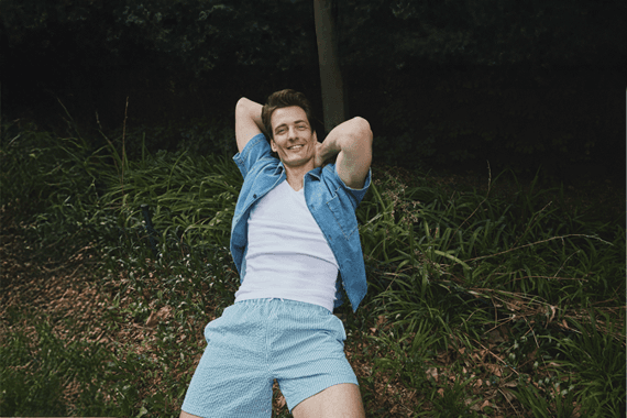 Mann in kurzärmeliger Hemdjacke und Shorts liegt mit verschränkten Armen im Gras im Grünen