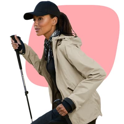 Frau in Outdoorjacke und Basecap wandert mit Stöcken vor rosa Hintergrund