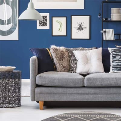 Graues Sofa mit gemusterten Kissen vor blauer Wand mit Kunstbildern, daneben schwarzes Regal und gemusterter Beistelltisch